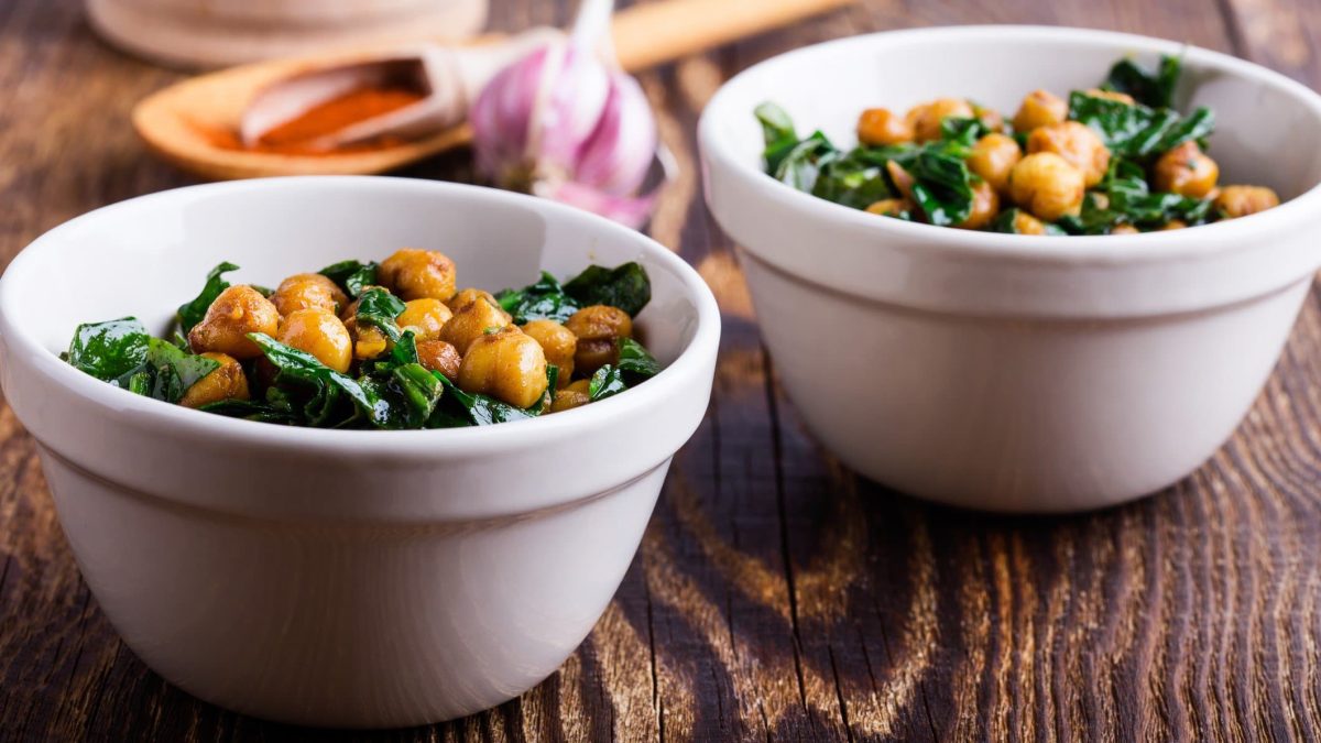 Zwei Schüsseln mit Nivik, ein Salat aus Kichererbsen und Spinat, stehen auf einem Holztisch. Im Hintergrund sind unscharf Knoblauch und ein Holzlöffel erkennbar.