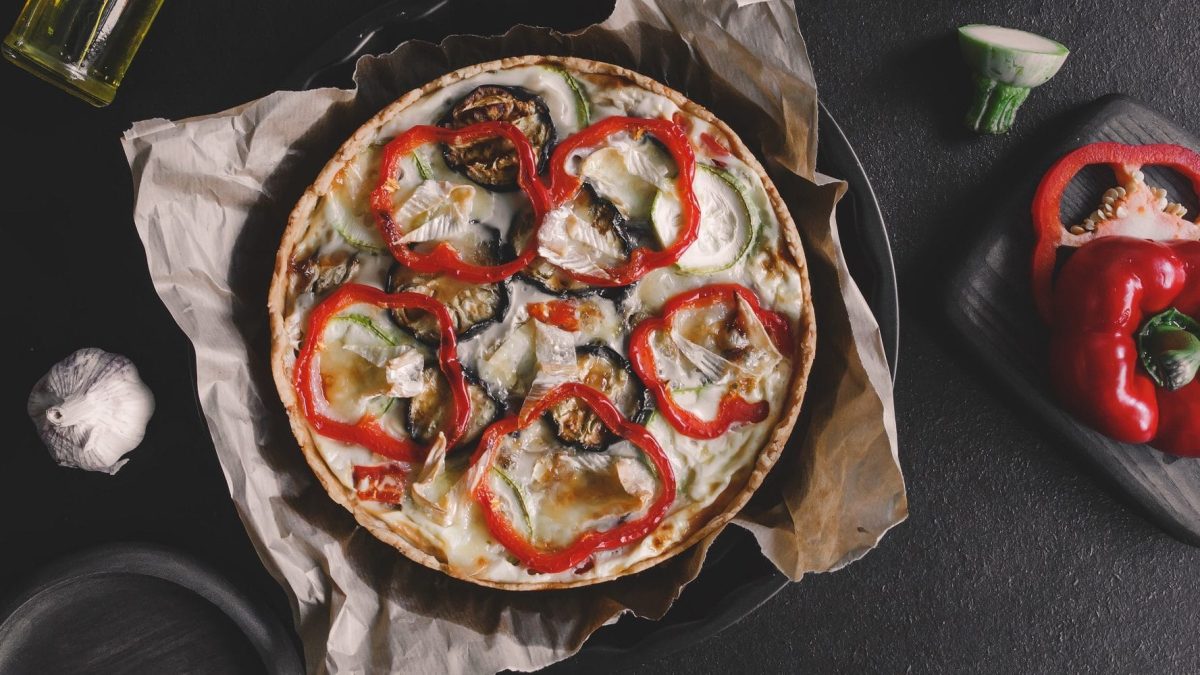 Draufsicht von oben: Auf einem Tisch liegt eine runde Form. In der ist eine rot weiße Füllung. Es handelt sich um eine Paprika-Ziegenkaese-Tarte.