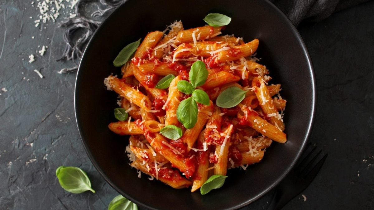 Ein schwarzer Teller mit Penne-Nudeln mit Tomatensauce, Parmesan und Basilikum steht auf schwarzem Hintergrund.