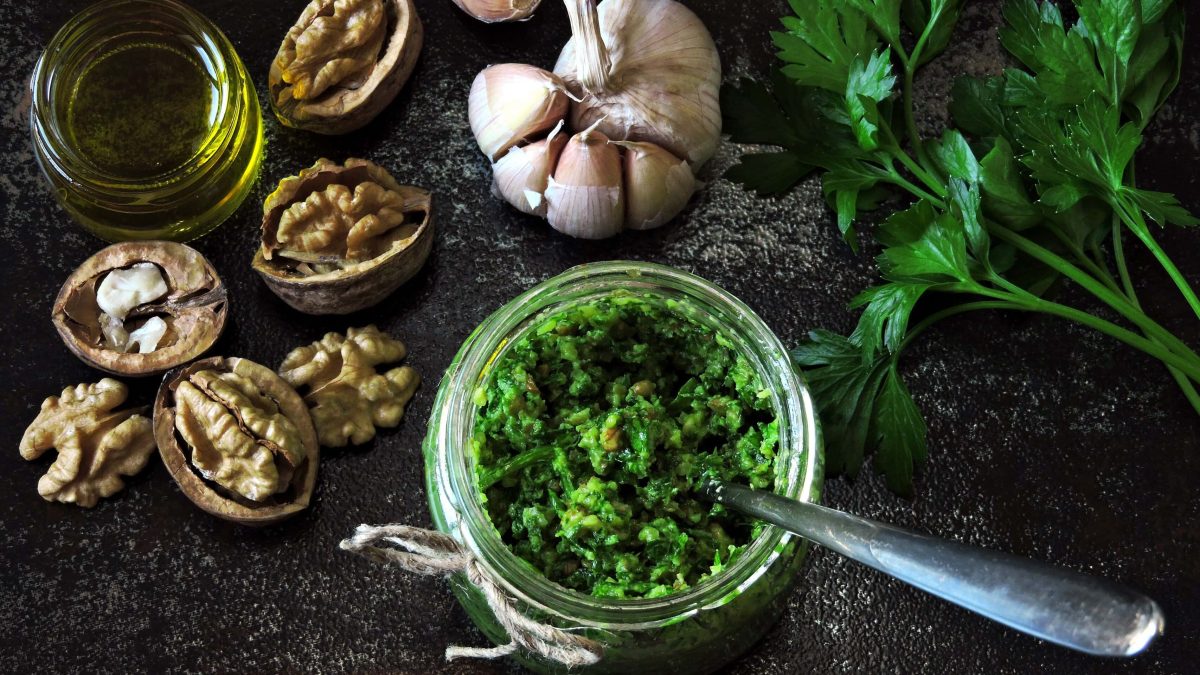 Draufsicht von oben: Auf einem schwarzen Hintergrund sind ein Knoblauch, Walnusshälften, Öl und Petersilienzweige zu sehen. Und in einem Gläschen ist Petersilien-Pesto mit Walnüssen abgefüllt.