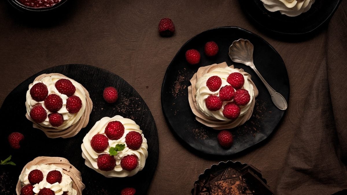 Mehrere schwarze Teller auf denen die Raspberry Meringue inspiriert von Downton Abbey angerichtet sind, neben einem Löffel, weiteren Himbeeren und einer Schale mit Schokolade, vor einem braunen Hintergrund, von oben.