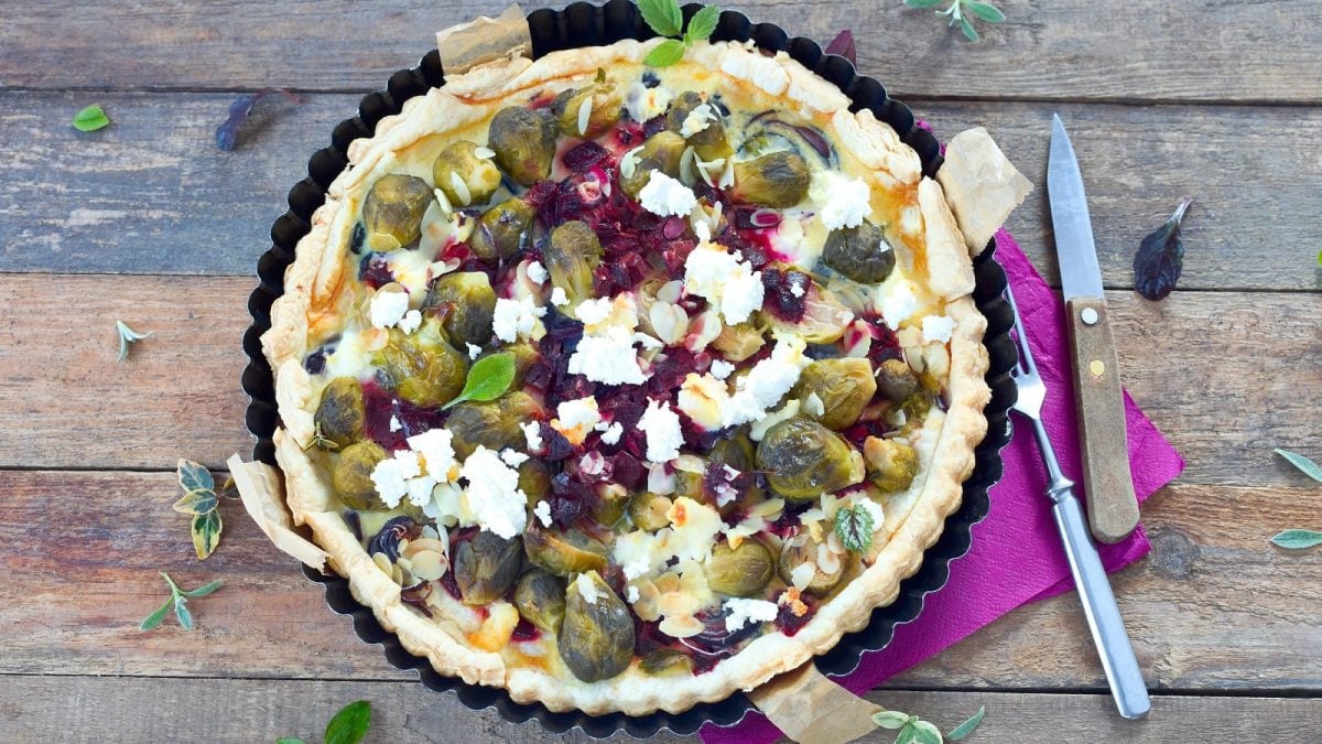 Eine Form mit der frisch gebackenen Rosenkohl-Rote-Bete Quiche mit Ziegenkäse, die mit Mandelblättchen und Kräutern garniert ist und neben der Besteck und eine lila Servierte liegt, vor einem Holzhintergrund, von oben.