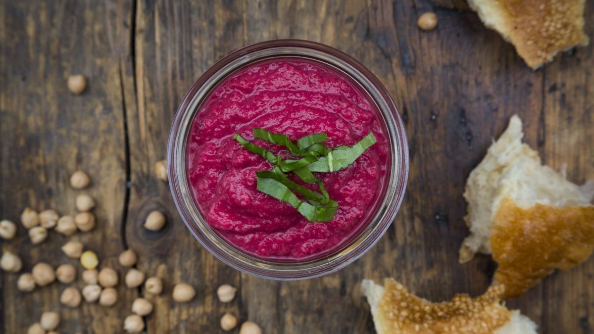 Auf einem rustikalen Tisch steht eine Schale mit Rote-Bete-Hummus. Daneben liegen Kichererbsen und Brot.