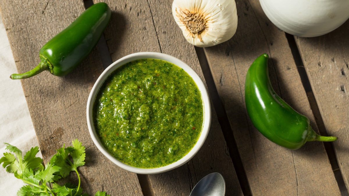 Auf einer Holzplatte liegen zwei grüne Chilischoten, ein Zweig Koriander und Knoblauch. Und eine weiße Schüssel in der eine grüne Paste zu sehen ist. Es handelt sich um die jemenitische Gewürzpaste Zhug.
