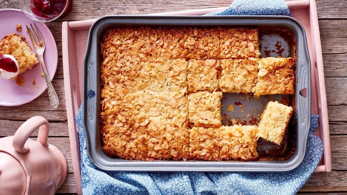 Schneller Butterkuchen in einer tiefen Kuchenform auf einem blau gemusterten Schirrtuch. Die Form steht auf einem rosanen Tablett auf einem hellbraunen Untergrund. Um die Form herum steht eine rosane Teekanne und ein Teller mit einem Stück Kuchen