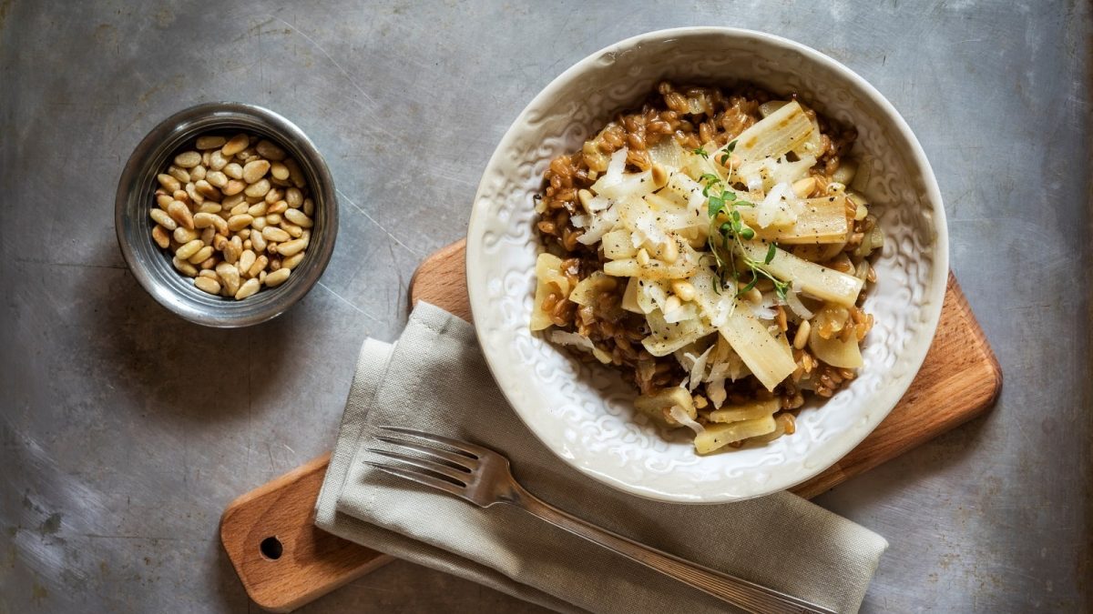 Eine Schale mit einer Portion Schwarzwurzel-Risotto garniert mit Pinienkernen, Parmesan und Kresse auf einem Holzbrett neben einer Stoff-Servierte, einer Gabel und einer Schüssel mit gerösteten Pinienkernen vor einem grauem Hintergrund, von oben.