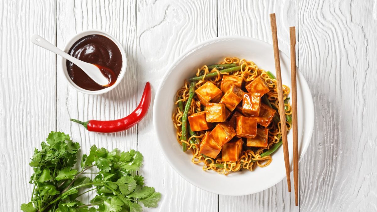 Auf hellem Holz steht in der Draufsicht ein Teller mit Ramennudeln und Tofu, daneben eine Schale selbst gemachter Hoisinsoße.