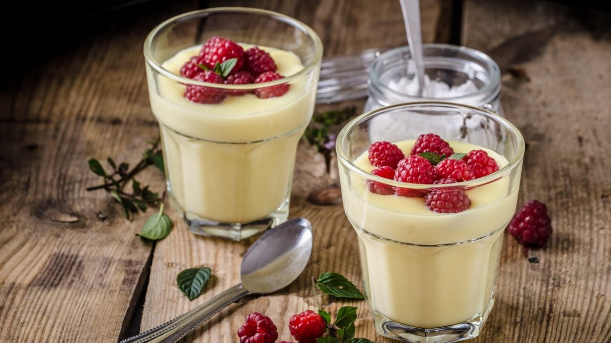 Zwei Gläsern mit Vanillepudding stehen auf einem Tisch. Auf dem Vanillepudding sind Himbeeren und Minzblätter. Auf dem Zisch liegt ein Löffel. Sowie ein flaches Glas mit einem weißem Puder.