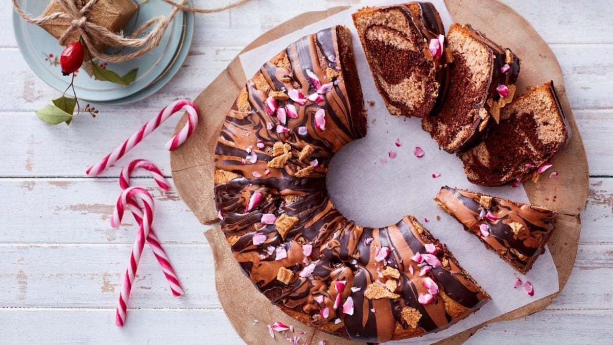 spekulatius-Mamorkuchen angeschnitten auf einem Holzbrett. Der Kuchen ist angerichtet mit Zuckerstangen und Spekulatius-Krümeln. Das Holzbrett steht auf einem weißen Holztisch.