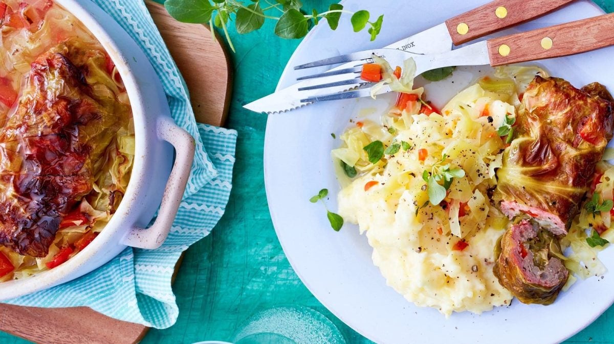 Ein Teller mit einer Portion Spitzkohl-Rouladen mit Gemüse auf Kartoffel-Püree neben Besteck, einem Bräter gefüllt mit weiteren Rouladen, der auf einem Holzbrett steht, und frischem Oregano, vor einem blauen Hintergrund, von oben.