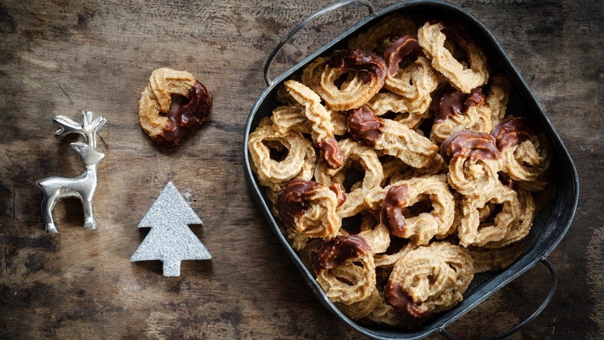 Eine Keksdose mit Spritzgebäck auf dunklem Holz in der Draufsicht, daneben liebt etwas Weihnachtsdeko.