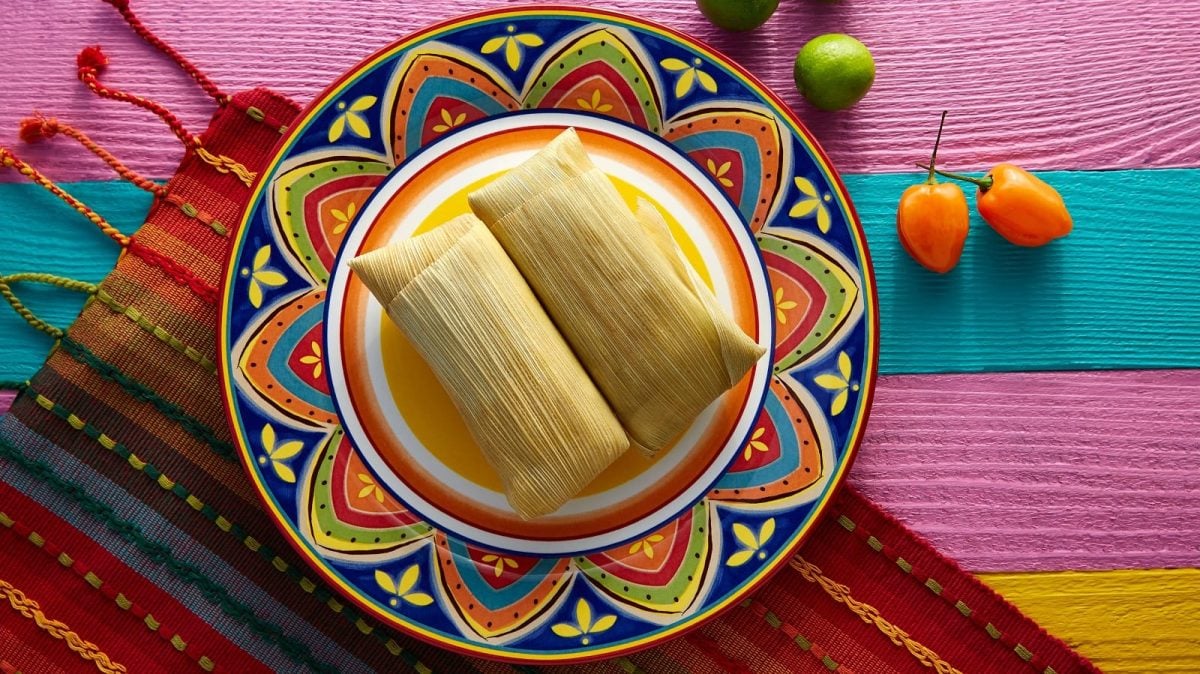 Zwei Tamales mit Schweinefleisch-Füllung auf einem bunt-verzierten Teller vor einem farbigen Teppich und Holzhintergrund neben Paprikaschoten und Limetten, von oben.