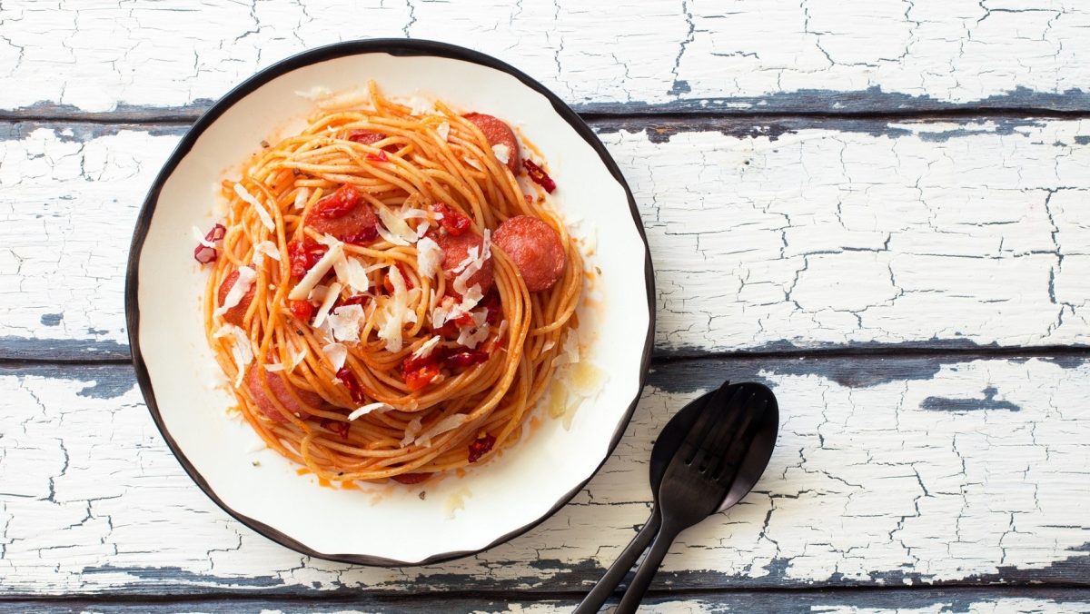 The Big Bang Theory Spaghetti mit Würstchen auf einem weißen Teller mit schwarzem Rand. Der Teller steht auf einem weißen Holztisch. Neben dem Teller liegen ein schwarzer Löffel und eine schwarze Gabel.