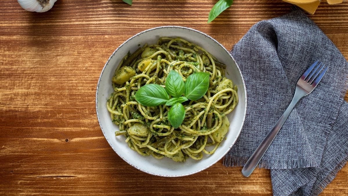 Eine Schüssel mit den Trenette al Pesto gefüllt und garniert mit Basilikumblättern, neben einem grauen Küchentuch auf dem eine Gabel liegt, vor einem Holzhintergrund, von oben.