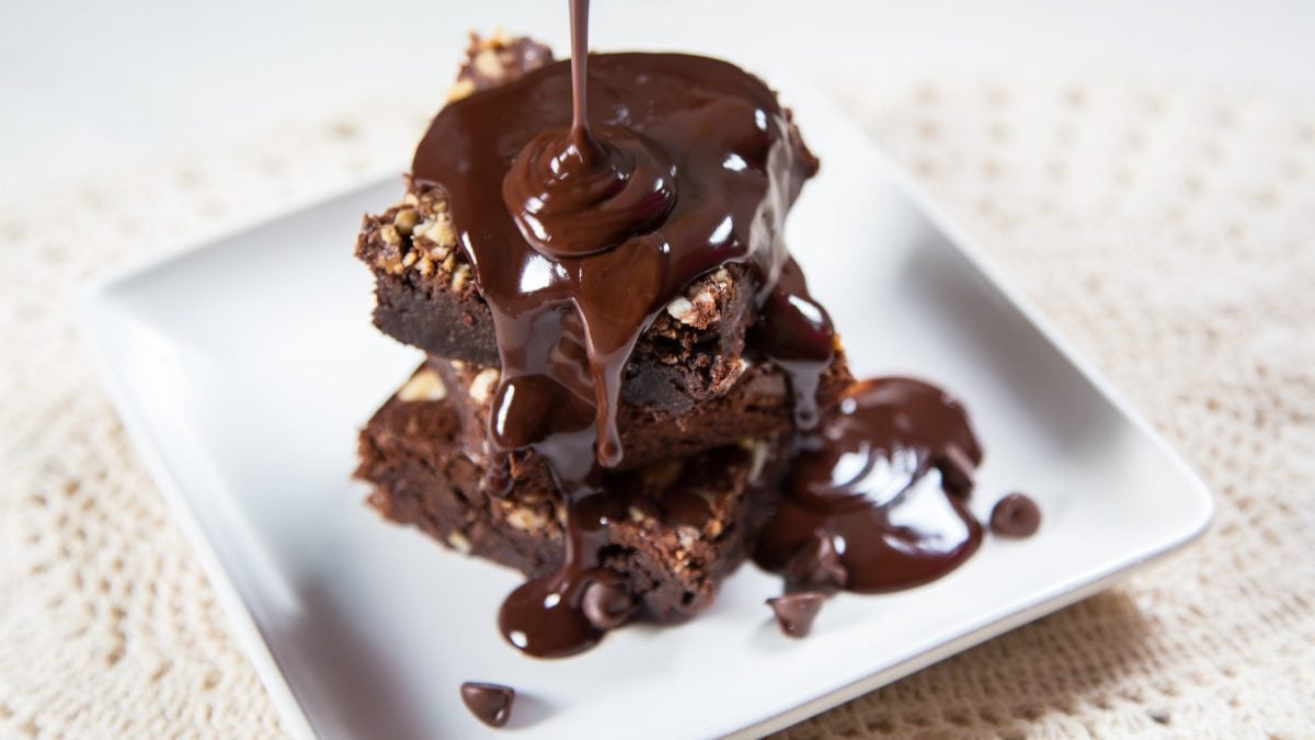 Triple Chocolate Brownie Stücke übereinander gelegt auf weißem Teller und beiger Tischdecke aus der Draufsicht im Moment des Übergießens mit dunkler Ganache.