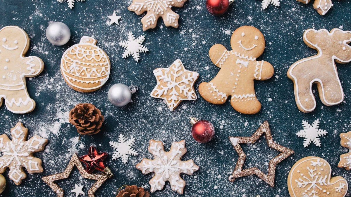 Auf dunklem Untergrun sind vegane, verzierte Pfefferkuchen zu sehen, außerdem Christbaumschmuck und Puderzucker.