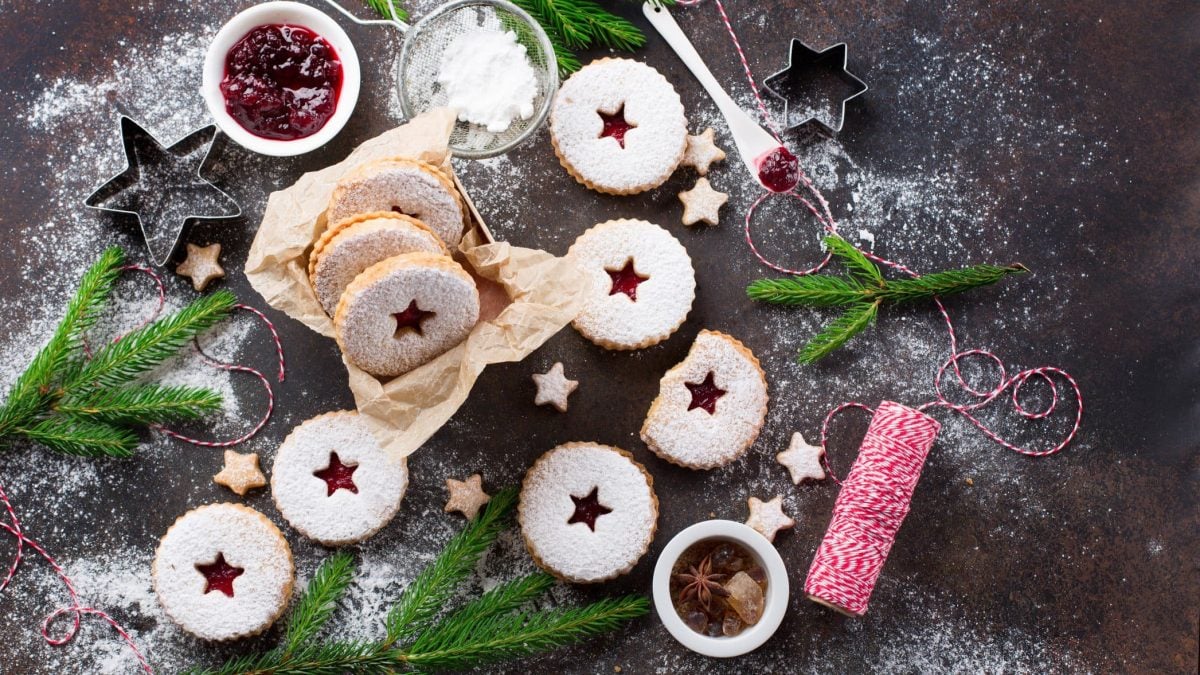 Vegane Spitzbuben auf dunklem Untergrund in der Draufsicht, bestäubt mit Puderzucker. Außerdem liegen ein paar Zutaten sowie weihnachtliche Deko herum.