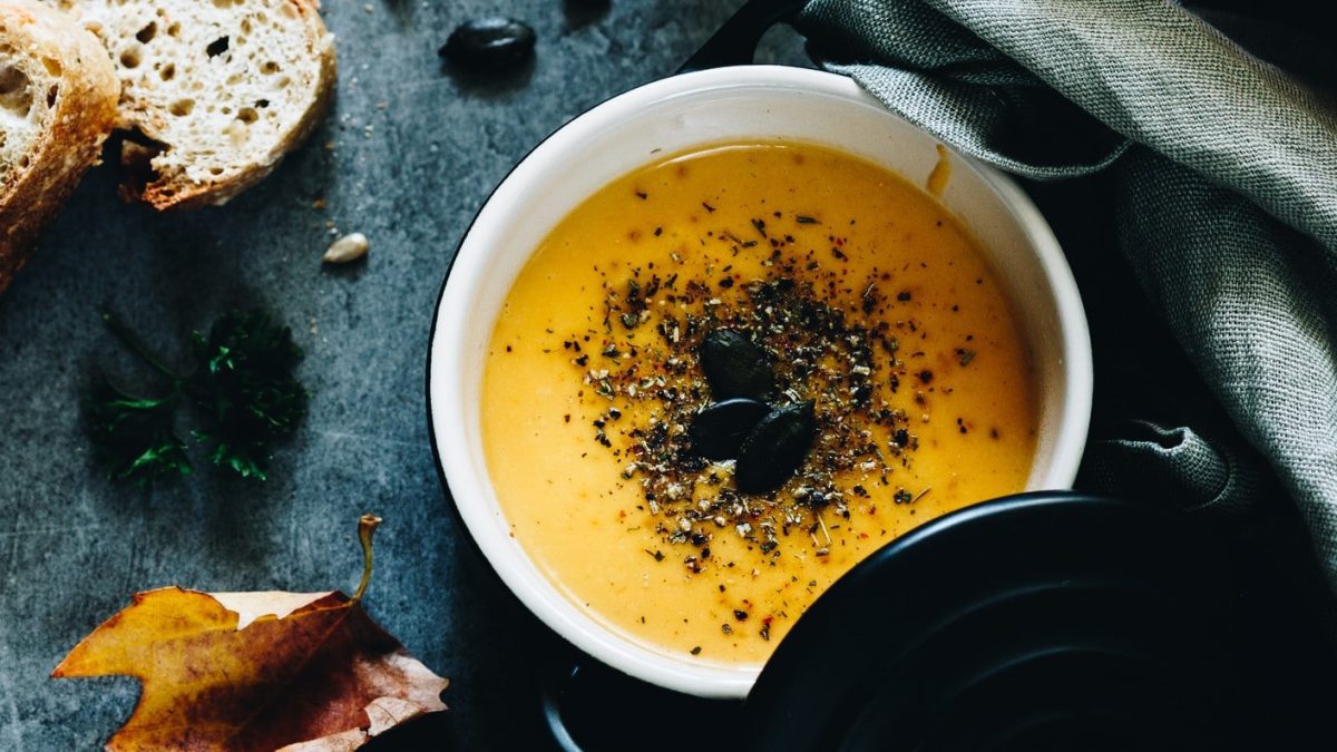 Ein Topf vegane Zucchini-Möhren-Suppe mit Kernen bestreut, daneben liegen zwei Scheiben Baguette und ien paar herbstliche Blätter. Alles in der Draufsicht.