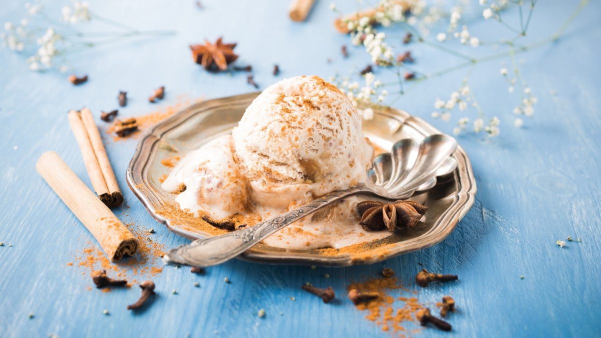 Eine Kugel veganes Zimteis liegt in einer kleinen Schale. Neben dem Eis liegt ein Löffel. Das Eis ist mit Zimt und Sternanis angerichtet. Der Teller steht auf einem blauen Untergrund. Um den Teller herum liegt Zimt, Sternanis und etwas Schleierkraut im Hintergrund