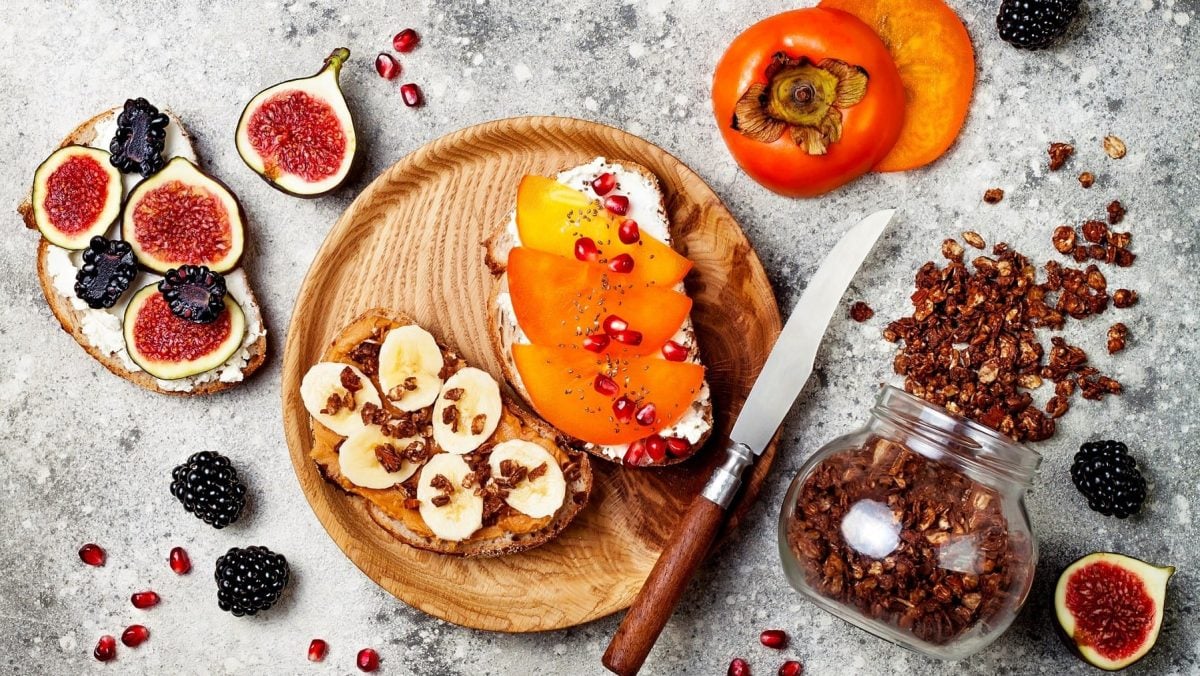 Frühstücksbrot auf einem runden Holzbrett auf einem grauen Untergrund. Ein Brot liegt links neben dem Brett und ist belegt mit Hüttenkäse, Feigen und Brombeeren. Die brote auf dem Brett sind angerichtet mit Erdnussbutter, Banane und Granola und Hüttenkäse, Kaki und Granatapfelkernen. Rechts neben dem Brett liegt ein umgekipptes Glas mit Granola.