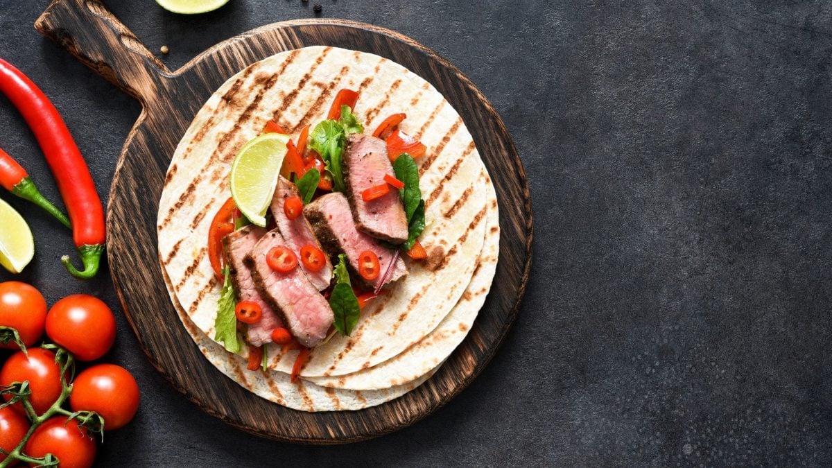 Auf einem runden Holzbrett liegen Wraps mit Roastbeef und Gemüse auf dunkel Untergrund, daneben sieht man Zutaten wie Tomaten und Chilis. In der Draufsicht.