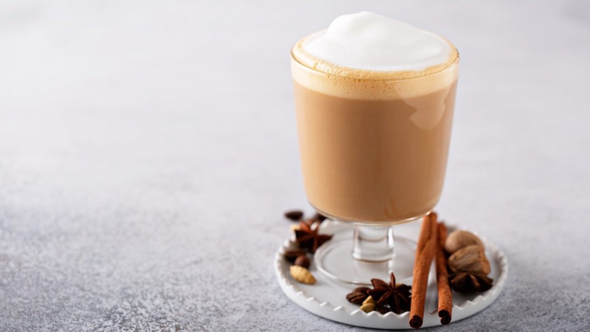 Ein Zimt-Chai-Latte in einem hohen Glas auf einem weißen Untersetzer. Der Untersetzer ist mit Nüssen, Zimtstangen und Anis dekoriert.