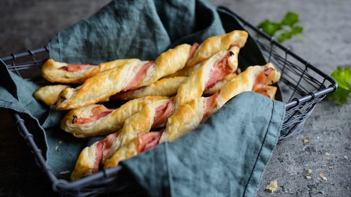 Gedrehte Blätterteigstangen mit Schinken in Brotkorb mit Geschirrtuch.