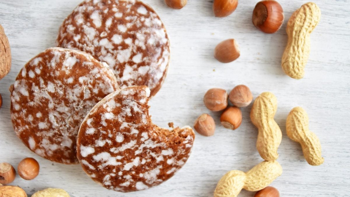 Mehrere Elisenlebkuchen auf einem hellen Untergrund aus Holz. Um die Lebkuchen herum liegen Haselnüsse und Erdnüsse. Einer der Lebkuchen ist bereits angebissen.
