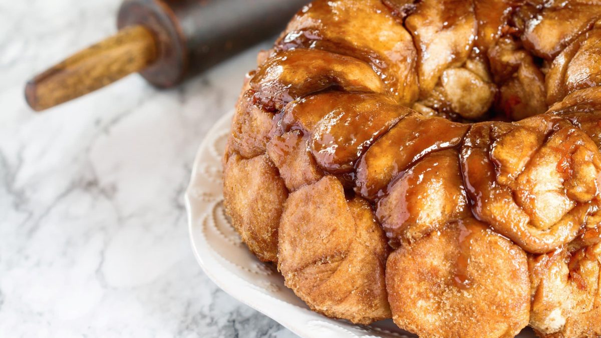 Süßes Monkey Bread mit Karamellsauce übergossen auf weißem Teller auf Marmorplatte. Am linken oberen Bildrand angedeutet schwarzes Nudelholz.