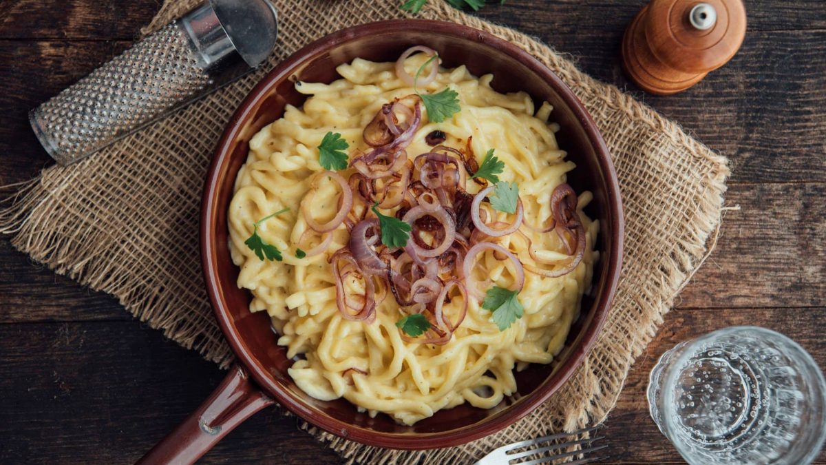 Pfanne mit Käsespätzle und Röstzwiebeln auf Holzuntergrund in rustikalem Ambiente.