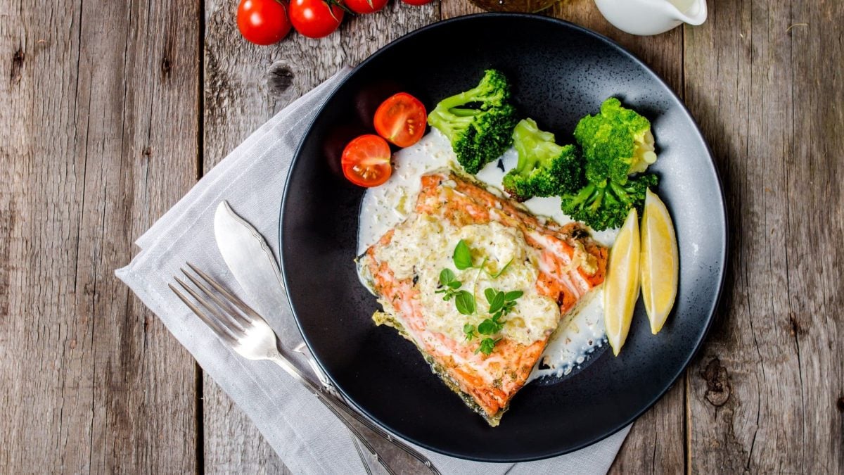 Lachs mit Weißweinsoße und Gemüse auf schwarzem Teller. Dieser steht auf hölzernem Untergurnd.