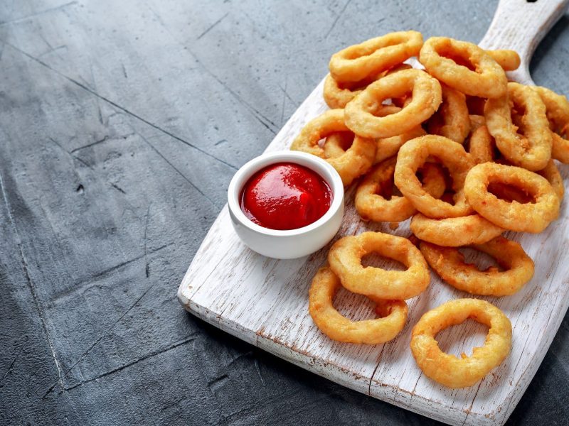 Onion Rings mit Ketchup auf Holzbrett, angerichtet auf grauem Untergrund