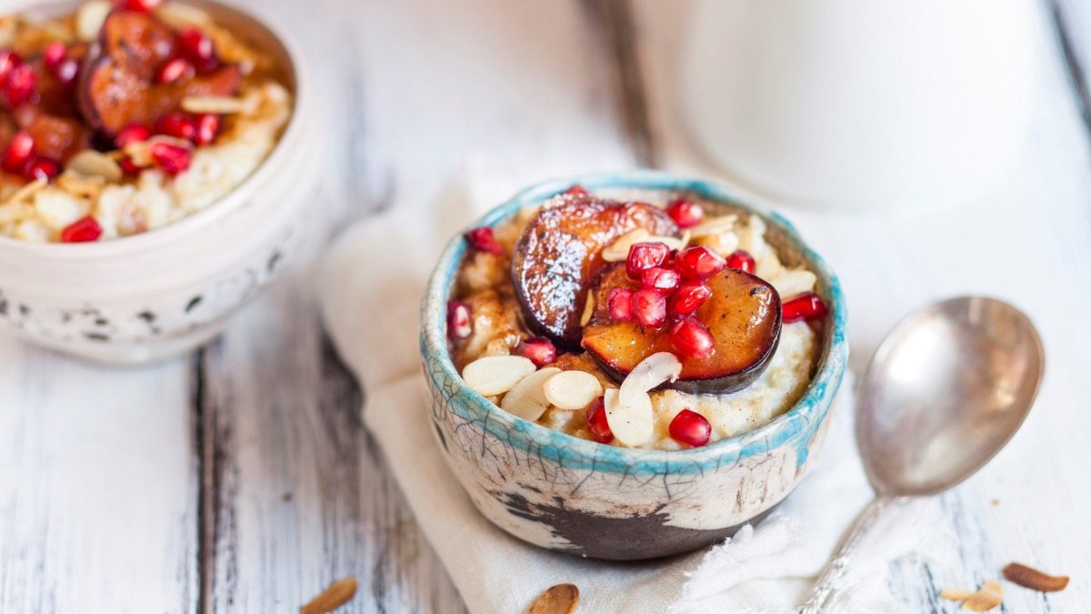 In einer Schüssel ist Pflaumen-Hafer-Bowl. Auf Haferbrei sind Pflaumen, Mandeln und Granatapfel. Daneben liegt ein Löffel.