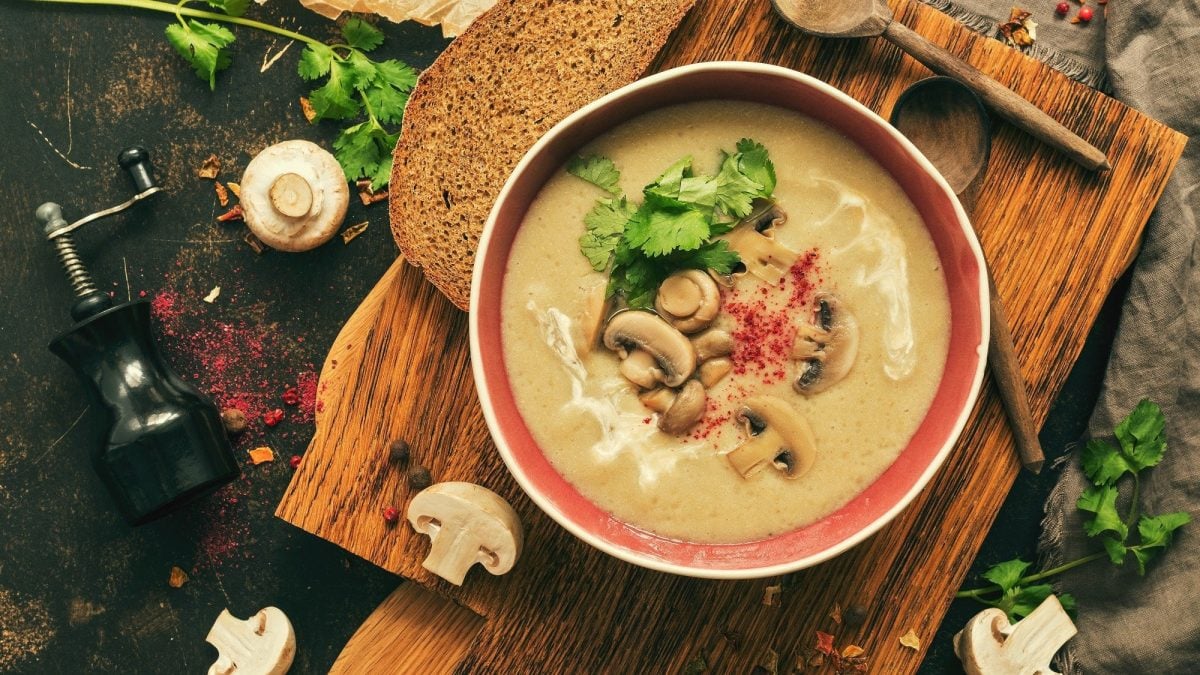 Eine Pilzcremesuppe mit gebratenen Pilzen, Chiliflocken und Petersilie steht auf einem dunklen Holzbrett in einer Schüssel. Um die Schüssel herum liegt Brot, Holzlöffel, Pilze, Petersilie und Gewürze.