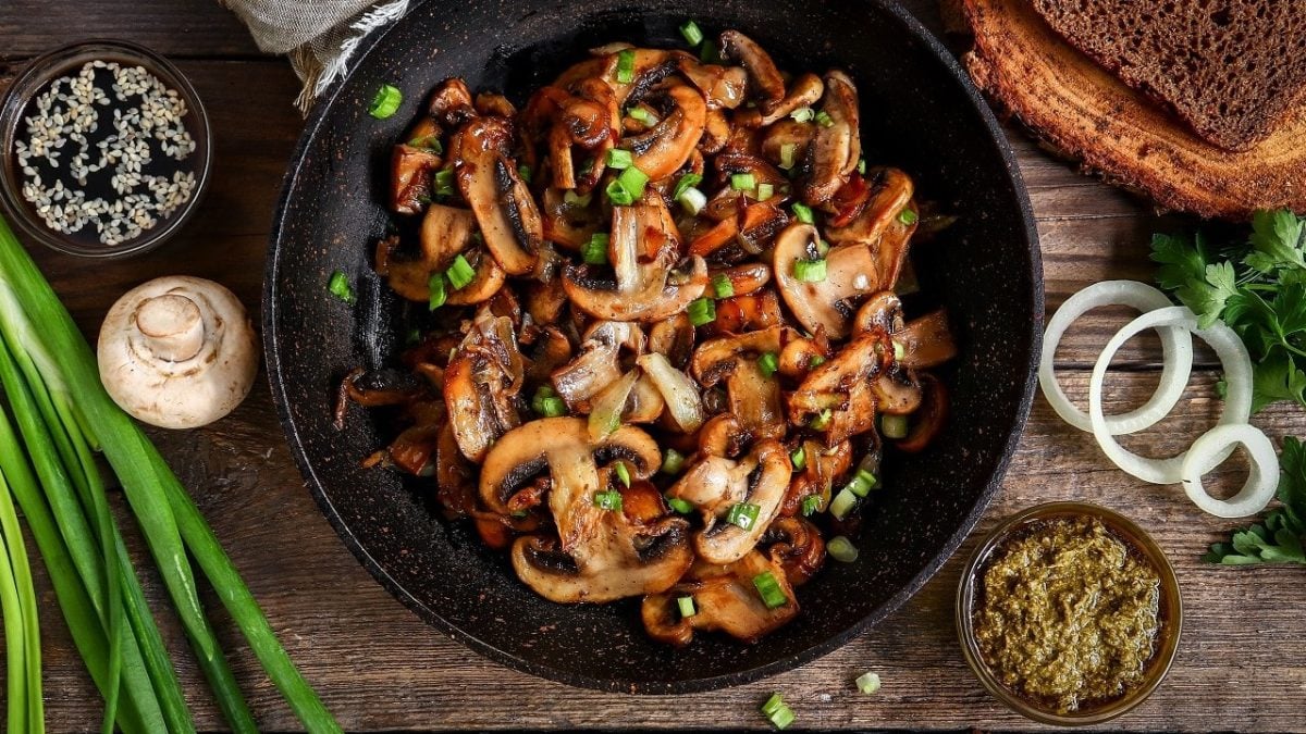 Gebratene Champignons mit Frühlingszwiebeln in Pfanne auf hölzernem Untergrund.