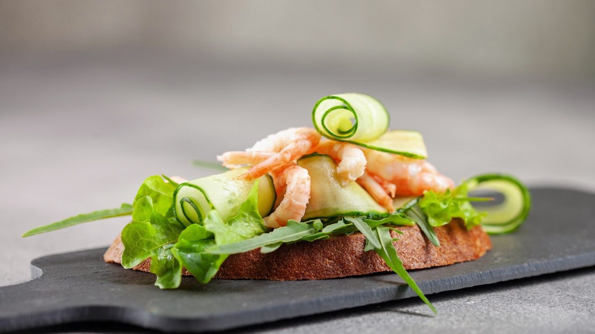 Sandwich mit Garnelen. Das Sandwich steht auf einer schwarzen Schiefernplatte auf einem grauen Untergrund. Das Sandwich ist belegt mit Salat, Gurken und Garnelen.
