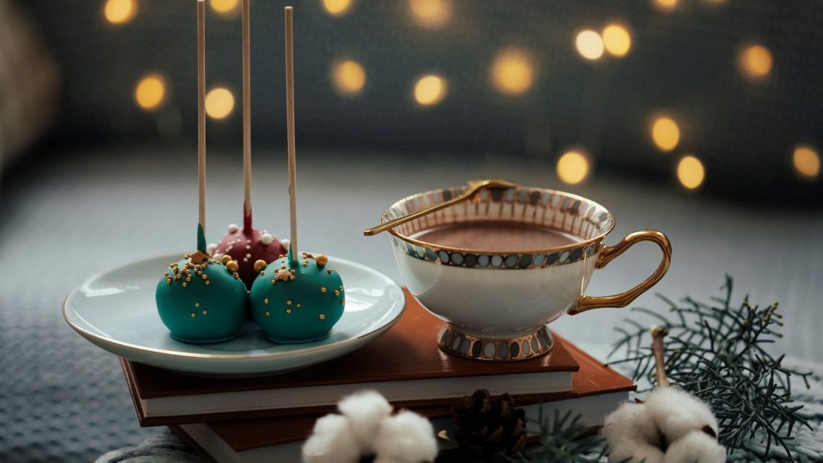 Auf einem Buch steht ein Teller mit weihnachtlichen Cake-Pops und eine Tasse Kakao. Die Cakepops stehen umgedreht auf dem Teller. Die Cake-Pops sind grün und rot gefärbt und mit Zuckerperlen dekoriert.