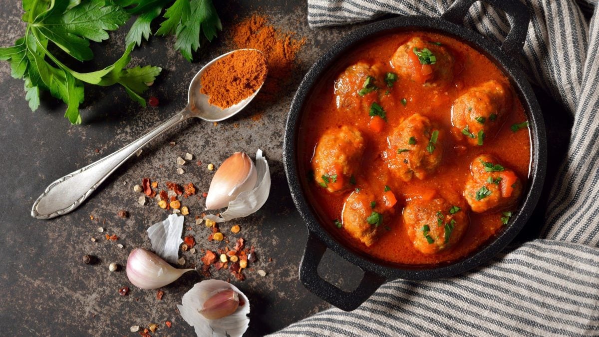Albóndigas con salsa de tomate in schwarzer Gussform mit Gewürzen und Teelöffel mit Paprikapulver aus der Vogelperspektive. Glatte Petersilie im linken oberen Bildrand. Recht dunkel-hellrau gestreifte Stoffserviette. Arrangiert auf dunklem Holz.