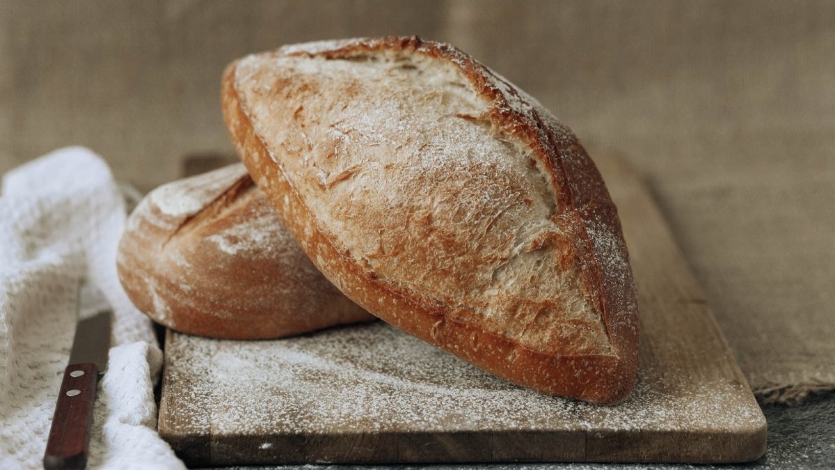 Zwei Baguette-Brötchen mit Kräuter der Provence auf einem Holzschneidebrett neben einem Brotmesser und einem weißen Tuch, vor einem braun-beigenen Hintergrund.