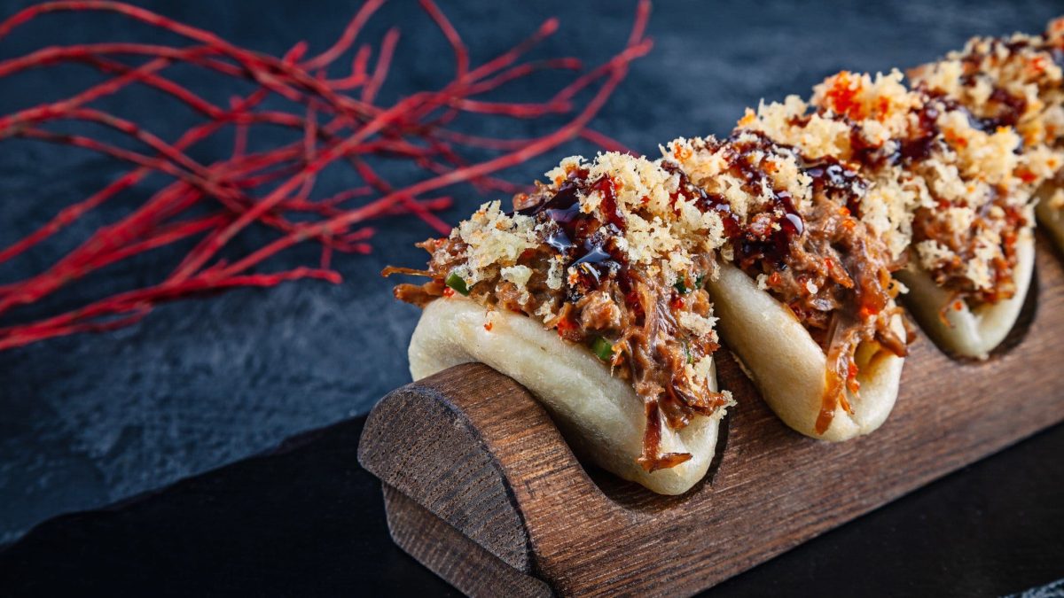 Vier Baos mit Char siu, Crunch und Hoisinsoße in Bambushalterung aus der Frontalsicht. Rote Chilifäden auf blauer Platte im Hintergrund.