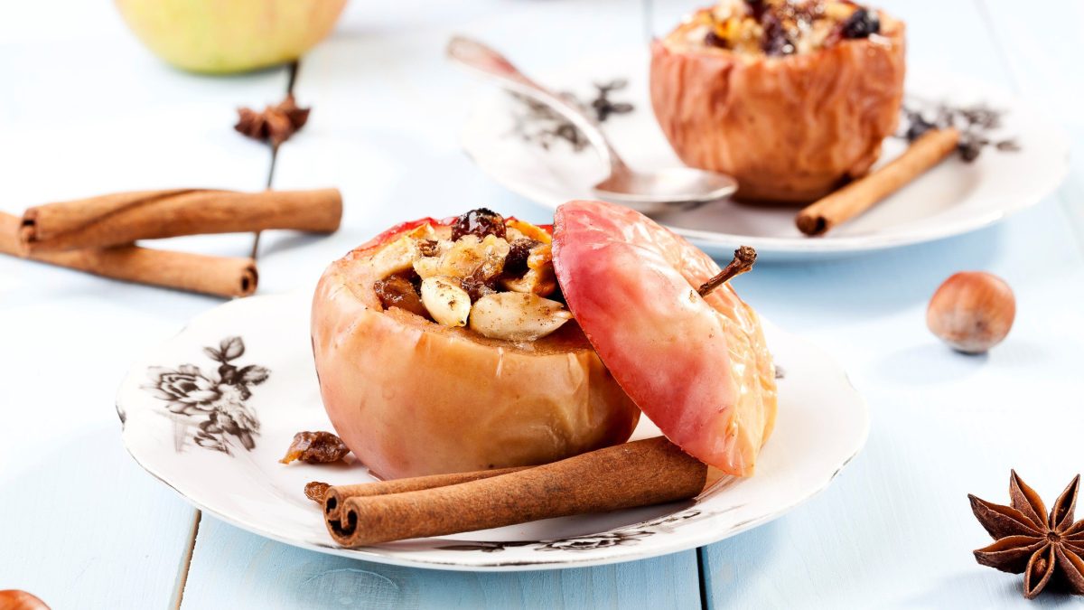 Zwei Bratäpfel mit Knusperhaube auf weißen Tellern, gefüllt mit Nüssen und Rosinen. Drumherum Zimtstangen, Nüsse, Anis.