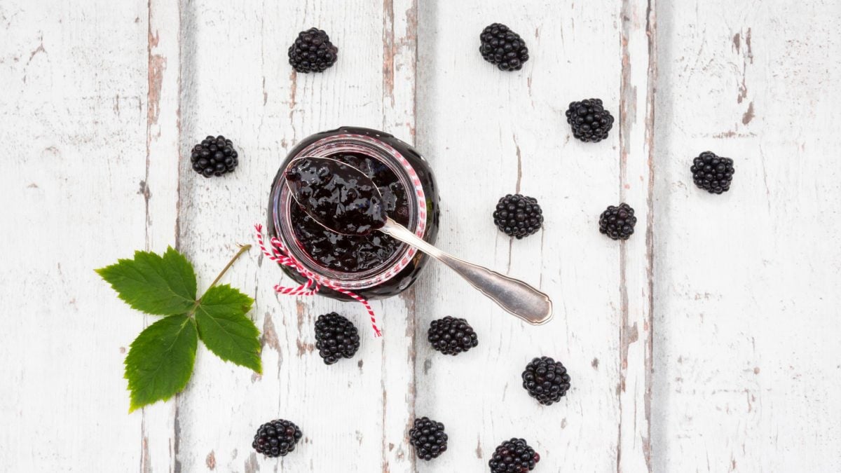 In der Draufsicht sieht man ein Glas Brombeermarmelade mit Chia auf hellem Holz, daneben liegen ein paar Brombeeren und Minze.