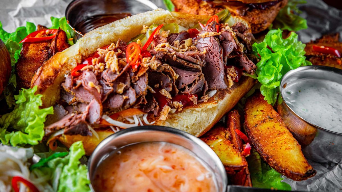 Cheese-Steak-Sandwich mit Soßen, Kartoffelecken und Salatdekoration mit Chiliringen und Röstzwiebeln aus der Frontalsicht.