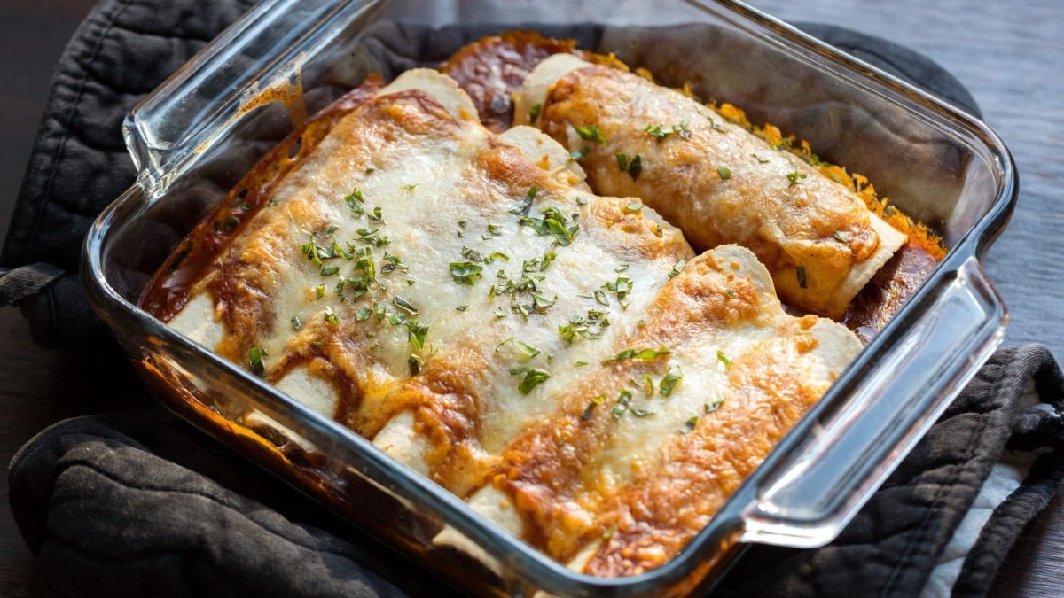Chili-Käse-Enchiladas in Auflaufform aus Glas auf grauem Topfhandschuh und dunklem Holztisch. Aufnahme aus der Draufsicht.
