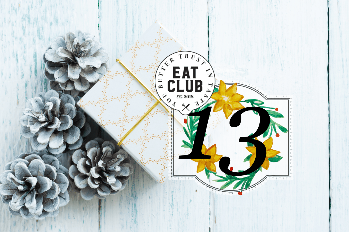 Ein weißes Paket verpackt und verziert mit goldenen Ornamenten neben weißen Tannenzapfen vor einem weißen Hintergrund, von oben, im Vordergrund ist eine 13 und das EATCLUB Logo zu sehen für das 13. Fenster des CLUBventskalenders mit dem Thema Weihnachtsessen in Spanien.