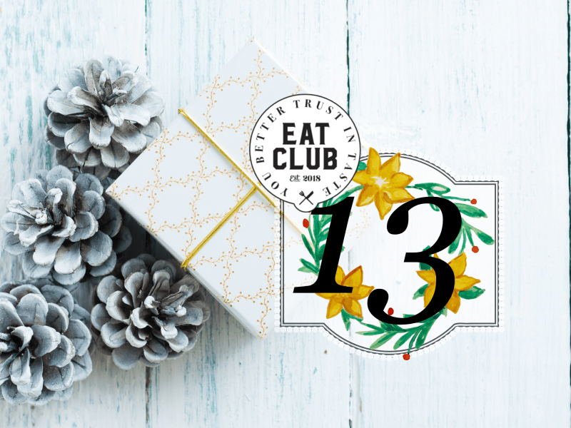 Ein weißes Paket verpackt und verziert mit goldenen Ornamenten neben weißen Tannenzapfen vor einem weißen Hintergrund, von oben, im Vordergrund ist eine 13 und das EATCLUB Logo zu sehen für das 13. Fenster des CLUBventskalenders mit dem Thema Weihnachtsessen in Spanien.