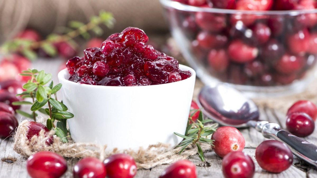 Auf einem Holztisch steht eine Cranberry-Marmelade. Die ist in einem weißen Schälchen. Drumherum liegen Cranberry. Auch eine GLasschüssel mit Cranberrys ist zu sehen.