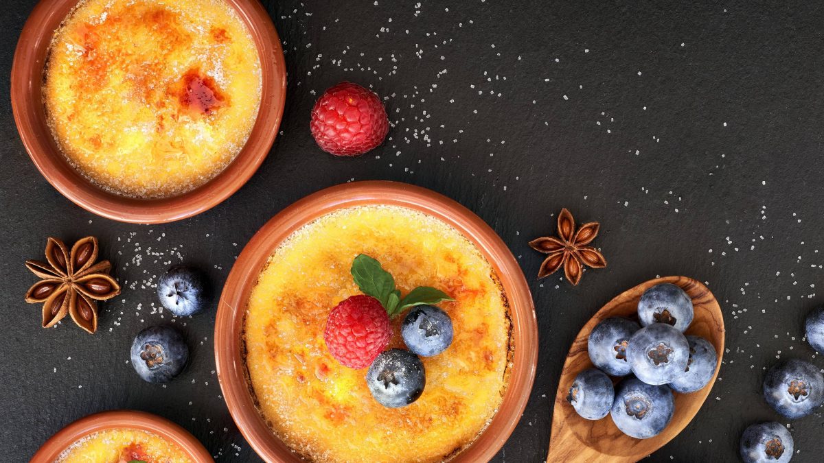Crema Catalana in Tonförmchen auf schwarzer Platte. Dekoriert mit Blaubeeren, Himbeeren, Minze und Sternanis. Aufnahme aus der Vogelperspektive