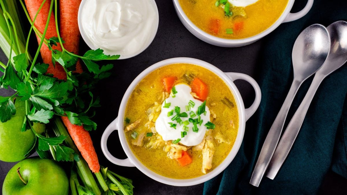 Zwei Mulligatawny Suppe aus Dinner for One in weißer Suppenschale mit zwei Henkeln auf schwarzer Schieferplatte. Auf der Suppe ein Kleks Creme Fraiche und Schnittlauch. Rechts unten zwei Suppenlöffel. Links Karotten, Petersilie und Apfel. Darüber eine kleine weiße Schale mit Creme Fraiche. Aufnahme aus der Vogelperspektive.