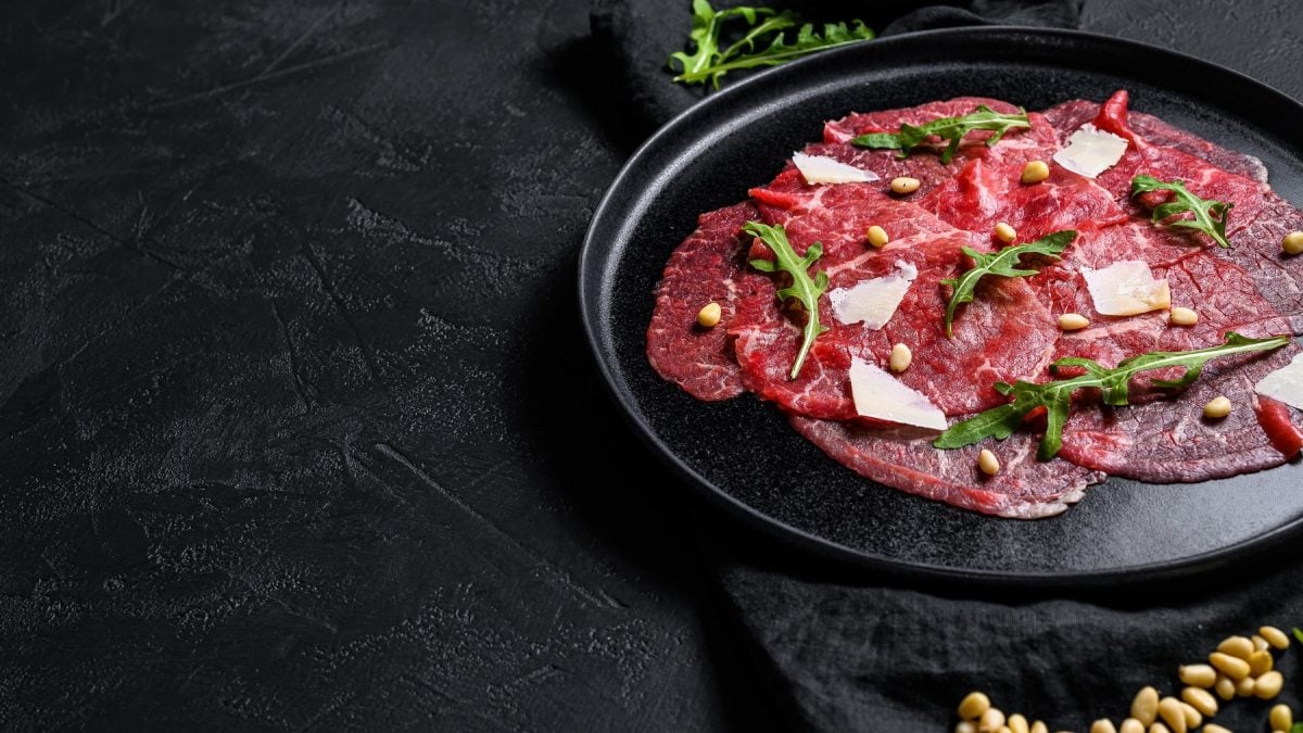 Blick von Oben: Auf einem schwarzen Hintergrund liegt ein Rinder-Carpaccio, das mit Parmesanhobeln, Pinienkernen und Rucola serviert wird.